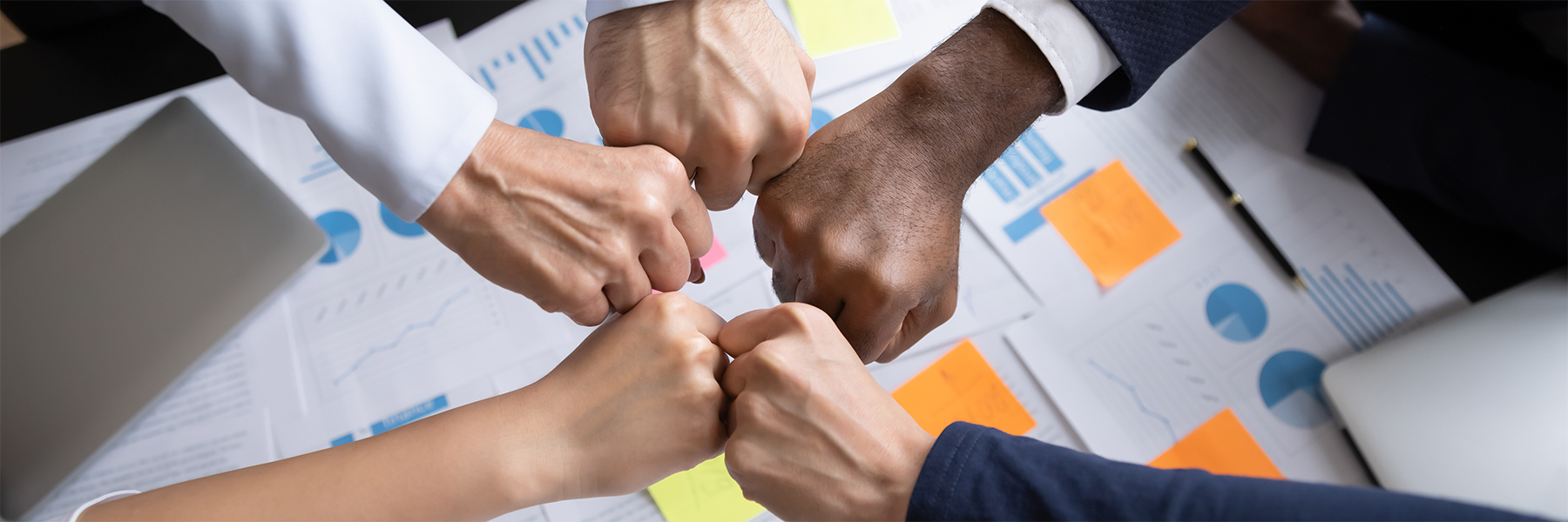 Close up of multiethnic business team bumping fists above the table with documents in office showing collaboration, support, unity, partnership, motivation and effective work together.
